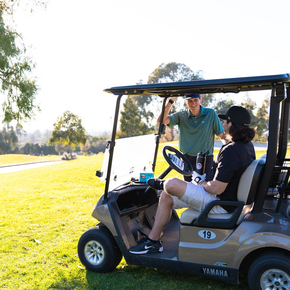 two men laughing in golf cart while drinking swing strong hydrate+ stick pack electrolyte energy hydration drink mix for golf